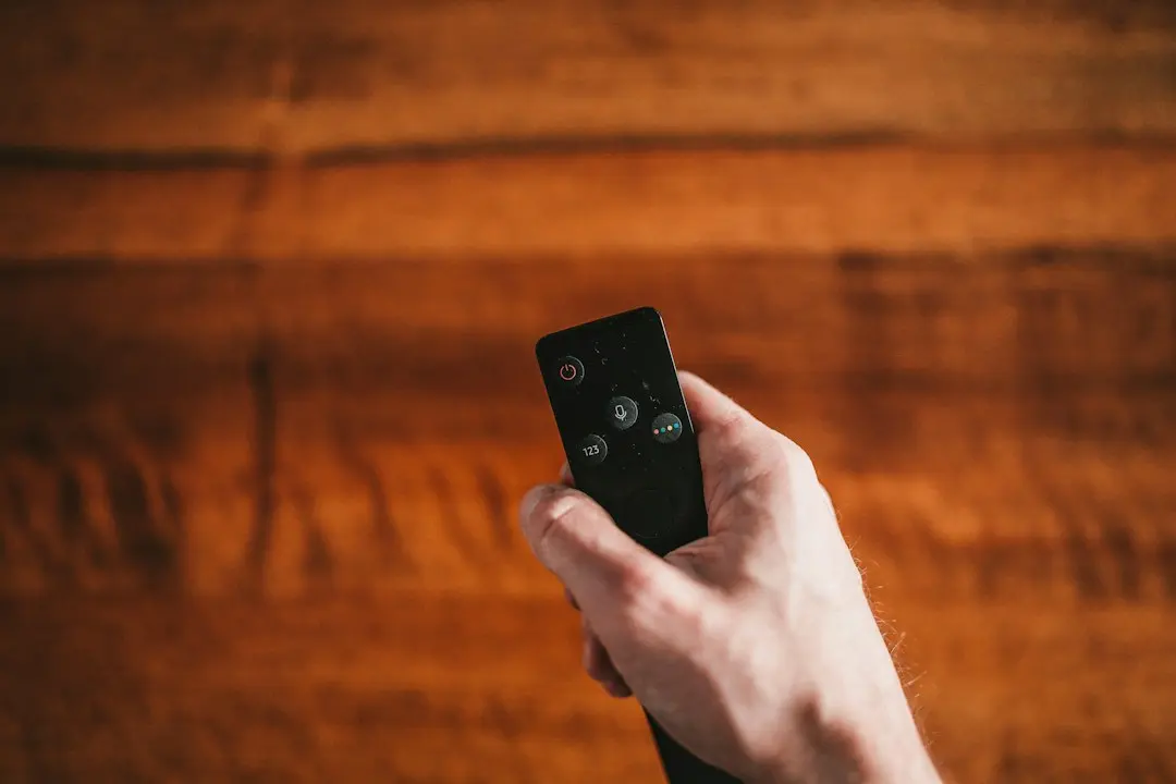 A hand holding a TV remote against a warm wooden background, about to choose the app to watch TvNado on